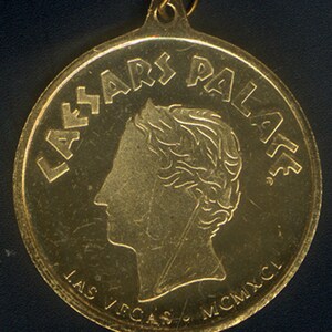 May include: A gold-colored metal token with the words "Caesar's Palace" and "Las Vegas MCMXCL" inscribed around the edge. A profile of a person's head is in the center.