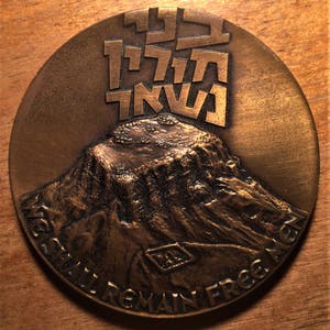 May include: A bronze medal with a mountain scene and the text "We Shall Remain Free" around the edge. The top of the medal features Hebrew text.