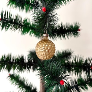 May include: A round, textured gold glass Christmas ornament with a bumpy, dimpled surface. The ornament features a metallic silver-toned cap at the top with a wire loop for hanging, suspended from a green artificial pine branch with red berries.