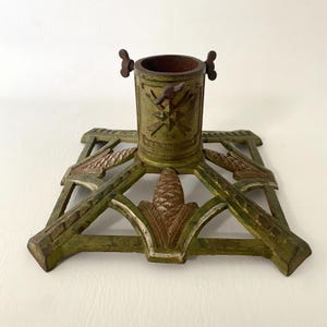 May include: An antique Christmas tree stand with a square base and a central cylindrical holder. The stand is painted in shades of green and brown, with decorative elements including a pinecone and stylized leaves. The stand has a height of approximately 10 inches.