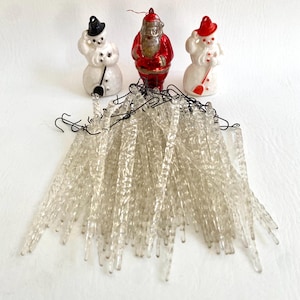 May include: A collection of vintage Christmas decorations, including three small figurines of a snowman, Santa Claus, and another snowman. A string of clear, icicle-shaped lights is in the foreground. The figurines are white, red, and black. The lights have black wires.