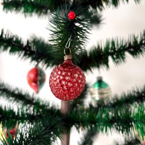 May include: A round, red Christmas ornament with a textured, dimpled surface hanging from a green artificial pine tree branch. The ornament features a metallic silver cap and a wire hook. Other blurred ornaments are visible in the background.