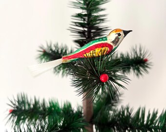 May include: A bird-shaped Christmas tree ornament featuring a painted body with green, red, and yellow accents, a pointed black beak, and a long, thin white tail made of fine bristles, positioned horizontally on a green artificial pine branch.