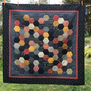 May include: A square quilt featuring a patchwork of hexagonal fabric pieces in various colours including mustard yellow, navy blue, red, and cream. The quilt has a denim-coloured inner border, a thin red accent border, and a dark navy outer border.