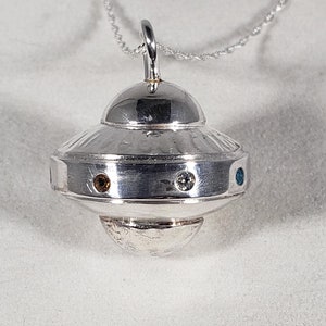 May include: A silver-toned pendant shaped like a flying saucer suspended from a delicate chain. The metallic craft features a domed top, a central band with three small circular inset stones in brown, clear, and blue, and a rounded bottom section.
