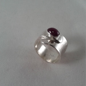 Puede incluir: Un anillo de plata con una banda texturizada y una piedra preciosa de color rojo oscuro engastada en un engaste de bisel. El anillo est&aacute; marcado con "Sterling".