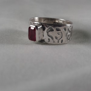 May include: A silver ring with a textured band and a red gemstone set in the center.