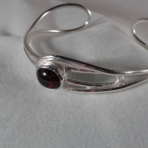 May include: A silver bangle bracelet with a red gemstone set in the center. The bracelet has a simple, elegant design with three bands of silver.