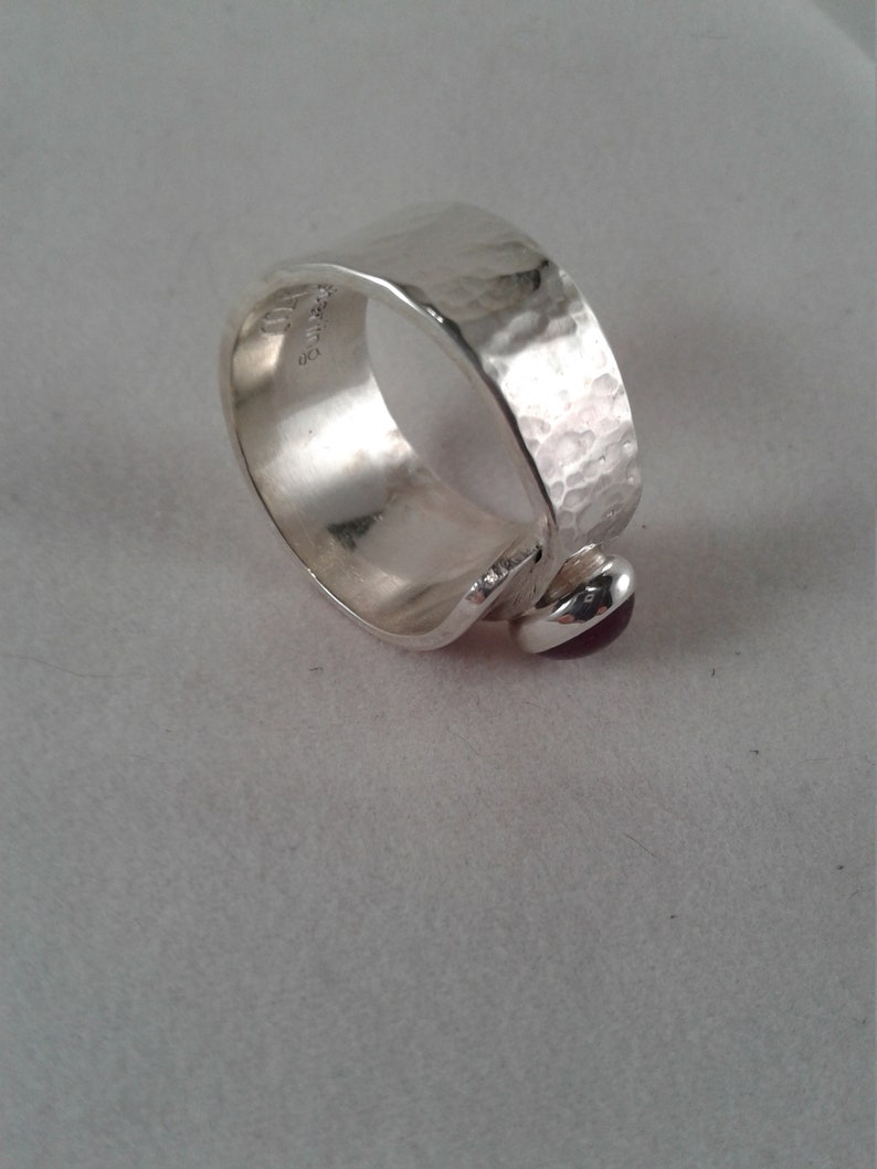 Puede incluir: Un anillo de plata con textura martillada y una peque&ntilde;a piedra preciosa p&uacute;rpura engastada en la banda.