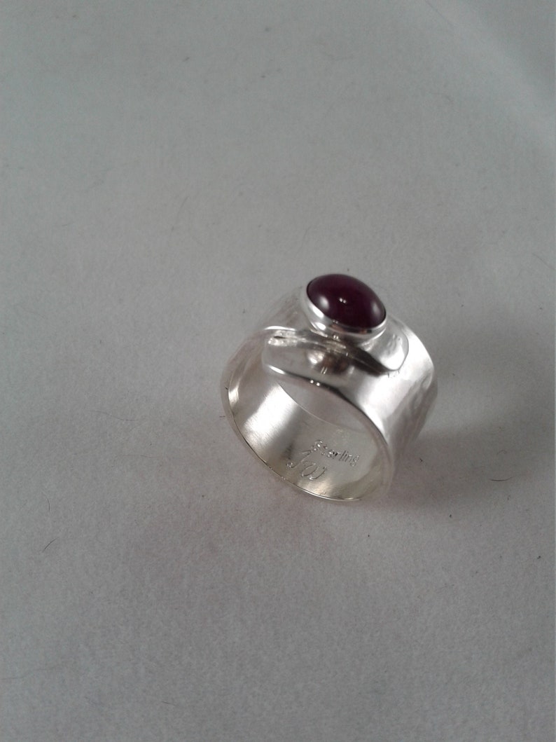 Puede incluir: Un anillo de plata con una banda texturizada y una piedra preciosa de color rojo oscuro engastada en un engaste de bisel. El anillo est&aacute; marcado con "Sterling".