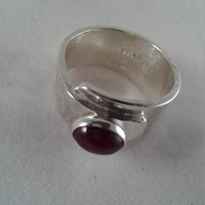 Puede incluir: Un anillo de plata con una piedra preciosa roja oscura engastada en un engaste de bisel. El anillo es ancho y tiene una banda texturizada.