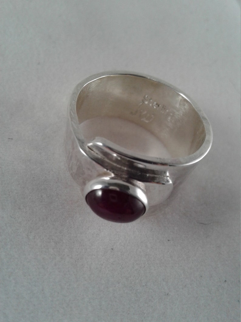 Puede incluir: Un anillo de plata con una piedra preciosa roja oscura engastada en un engaste de bisel. El anillo es ancho y tiene una banda texturizada.