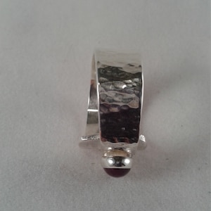 Puede incluir: Un anillo de plata con una banda martillada y una piedra preciosa de color rojo oscuro.