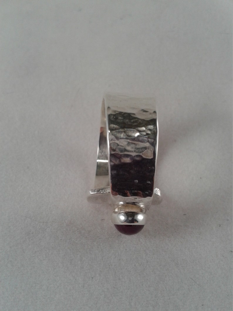 Puede incluir: Un anillo de plata con una banda martillada y una piedra preciosa de color rojo oscuro.