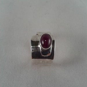 Puede incluir: Un anillo de plata con una piedra preciosa de color rojo oscuro engastada en el centro. El anillo tiene una banda texturizada y un dise&ntilde;o &uacute;nico.