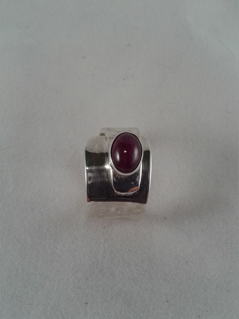Puede incluir: Un anillo de plata con una piedra preciosa de color rojo oscuro engastada en el centro. El anillo tiene una banda texturizada y un dise&ntilde;o &uacute;nico.