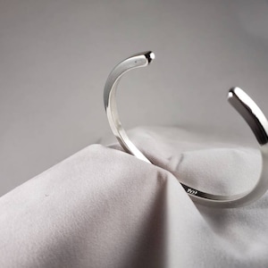 May include: A silver bangle bracelet with a smooth, rounded surface. The bracelet is open and has a slightly curved shape.