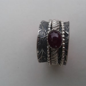 May include: A silver ring with a textured band and a deep red gemstone set in the center. The ring is made of three bands, each with a different design.