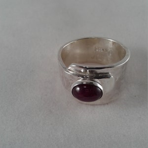 Puede incluir: Un anillo de plata con una piedra preciosa de color rojo oscuro engastada en un engaste de bisel. El anillo es ancho y tiene un acabado texturizado.