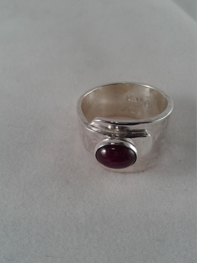 Puede incluir: Un anillo de plata con una piedra preciosa de color rojo oscuro engastada en un engaste de bisel. El anillo es ancho y tiene un acabado texturizado.