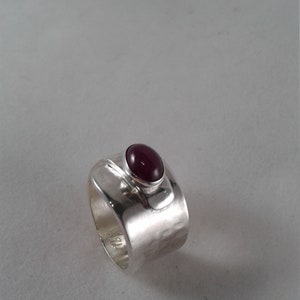 Puede incluir: Un anillo de plata con acabado martillado y una piedra ovalada de color morado oscuro engastada en el centro.