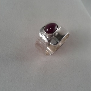 May include: A silver ring with a hammered texture and a deep red oval gemstone set in the centre.