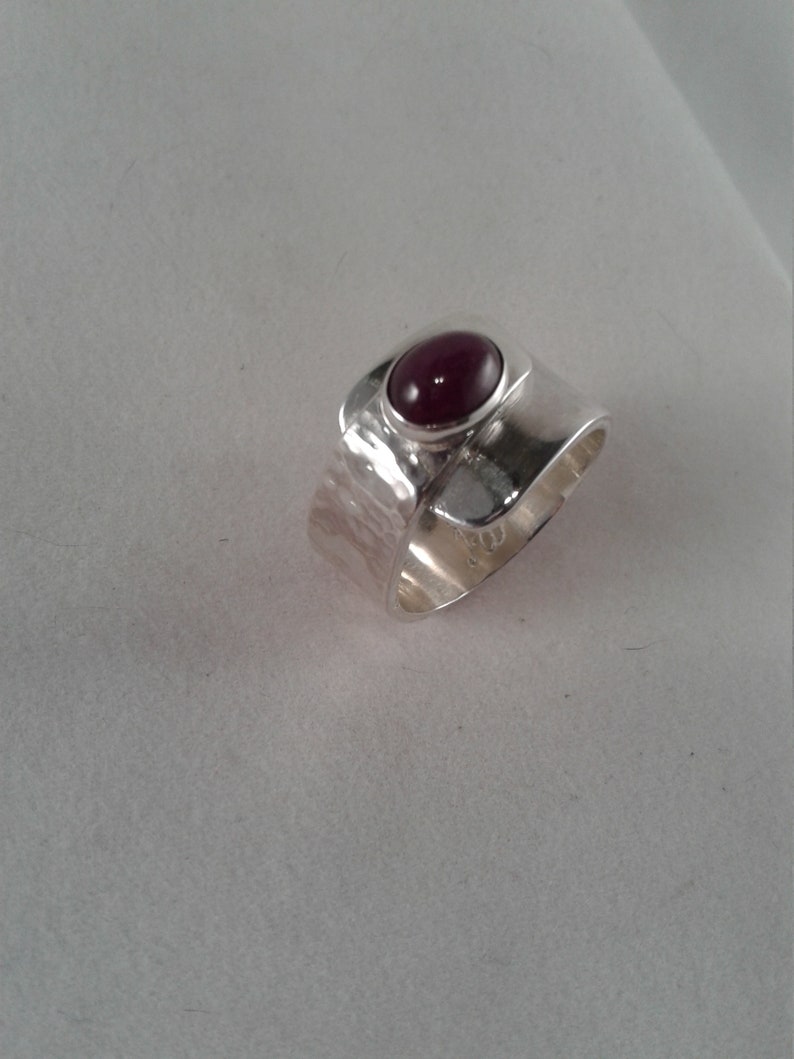Puede incluir: Un anillo de plata con textura martillada y una piedra ovalada de color rojo oscuro engastada en el centro.