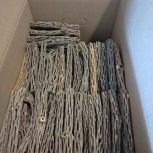 May include: A cardboard box filled with numerous pieces of dried cholla wood. The wood pieces are cylindrical, with a light gray color and a porous, open structure. The box is open, revealing the contents inside.