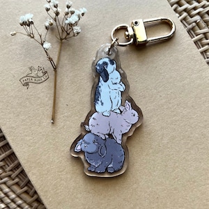 May include: A clear acrylic keychain featuring a stack of three cartoon rabbits in shades of gray, lavender, and black and white. The keychain has a gold-colored clasp and a small sprig of white flowers.