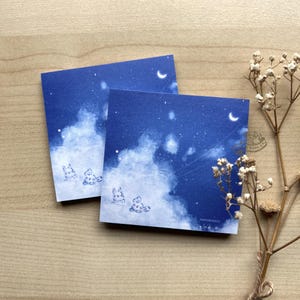 May include: Two square blue notepads with a night sky design featuring clouds, stars, and a crescent moon. The bottom of the notepads have a small illustration of a bunny. The notepads are on a wooden surface.