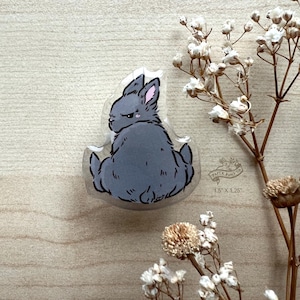 May include: A gray rabbit with pink ears sits on its hind legs with its back to the viewer. The rabbit is in a clear plastic button with the text "Paper & Pins Co. 1.5" x 1.25" printed on the bottom.