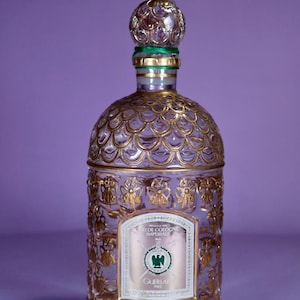 Op de afbeelding: Een heldere glazen parfumfles met gouden details en een decoratieve dop. De fles heeft een etiket met tekst en een groen embleem. Op een paarse achtergrond.