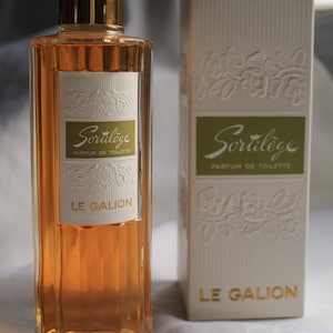 May include: A perfume bottle with a gold cap and amber liquid, alongside a matching box. The bottle and box display the text "Sortilège Parfum de Toilette LE GALION" in white and gold. The box has a floral embossed design.