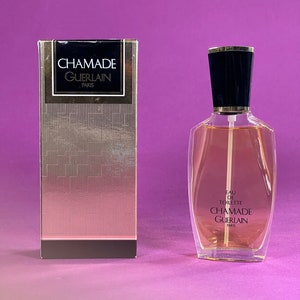 May include: A clear glass bottle of Chamade Eau de Toilette by Guerlain Paris. The bottle has a black cap and is sitting on a purple surface. The bottle is surrounded by a gold and black box with the same text.