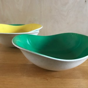 May include: Three ceramic bowls with a speckled off-white exterior and a smooth, colourful interior. One bowl has a yellow interior, while another has a green interior. The bowls have a unique, wavy shape, and are likely for serving.