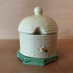 May include: A ceramic honey pot with a lid, in a light yellow hue and a textured, beehive-like pattern. The pot has a green base and a painted bee. The lid has a small opening for a spoon.