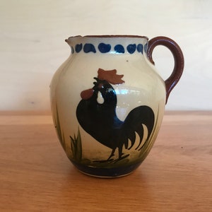 May include: A ceramic jug featuring a rooster design. The jug has an off-white body with a brown handle and rim. A black rooster is painted on the front, with green grass and blue accents. It is likely used for serving drinks.