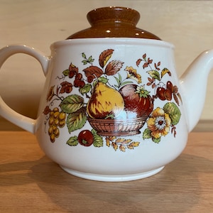May include: Cream ceramic teapot with a brown lid and handle. The teapot has a fruit basket design with pears, grapes, and apples in yellow, brown, and red. A classic kitchen item.
