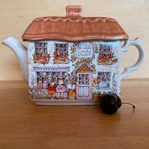 May include: A ceramic teapot in the shape of a cottage, with a brown roof and a facade decorated with windows, floral details, and the text "Ye Olde Candy Shoppe." A dark cherry sits on the wooden surface.
