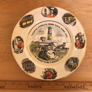 May include: A white plate with a gold rim, decorated with mining illustrations and workers. The plate features the text: "DESPITE PITFALLS, SOME GOOD, SOME BAD, I'M PROUD TO BE A MINING LAD."