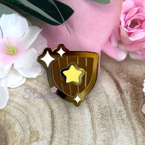 Puede incluir: Pin de esmalte de color dorado con un escudo con una estrella amarilla y destellos blancos. El escudo tiene una textura de madera.