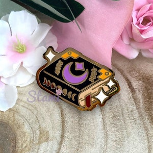 Peut inclure: Une épingle en émail dorée représentant un livre fermé avec une lune croissante violette et des étoiles sur la couverture. Le livre a une reliure dorée et des pages. L'épingle a un effet brillant.