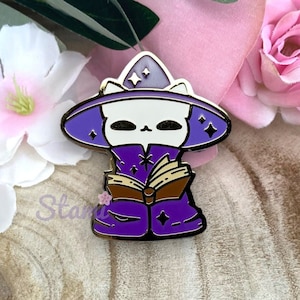 Op de afbeelding: Een witte kat die een paarse tovenaarsmantel en een puntige hoed met gouden sterren draagt. De kat houdt een boek in zijn poten. De pin is gemaakt van goudkleurig metaal met emaille accenten.