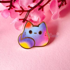May include: A colourful cat enamel pin with a gold border. The cat is a mix of blue, purple, orange, and yellow. The pin has a cute, cartoonish design. The pin is on a pink background with pink flowers.