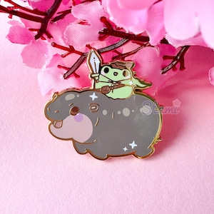 May include: An enamel pin depicting a grey hippo with a light pink belly and a green frog warrior riding on its back. The pin has a gold border and white star accents. The frog holds a sword. The pin is set against a pink background.