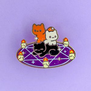 May include: Enamel pin showing three cartoon cats on a purple pentagram with candles. The cats are orange, white, and black. The pentagram has a gold border and is surrounded by lit candles.