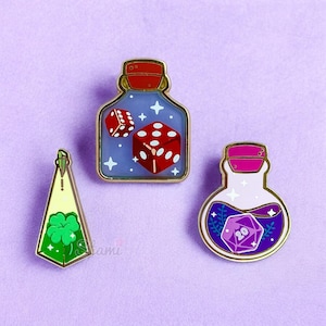 May include: Three enamel pins featuring glass bottles with different coloured liquids and dice inside. The first bottle is blue with a red and white die, the second is green with a four-leaf clover, and the third is purple with a 20-sided die.