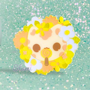 May include: A yellow enamel pin featuring a cartoon cat with a lollipop in its mouth. The cat is surrounded by yellow, orange, and white flowers.