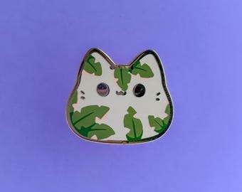 May include: A cat-shaped enamel pin with a white base and a pattern of green leaves. The pin has a gold-coloured outline and features black eyes and a small, black nose. The pin is set against a purple background.
