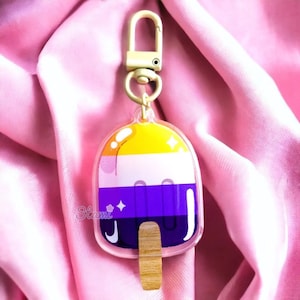 May include: A clear acrylic keychain shaped like a popsicle with the colors of the non-binary pride flag: yellow, white, purple, and dark purple. The popsicle has a wooden stick. The keychain has a gold metal clasp.